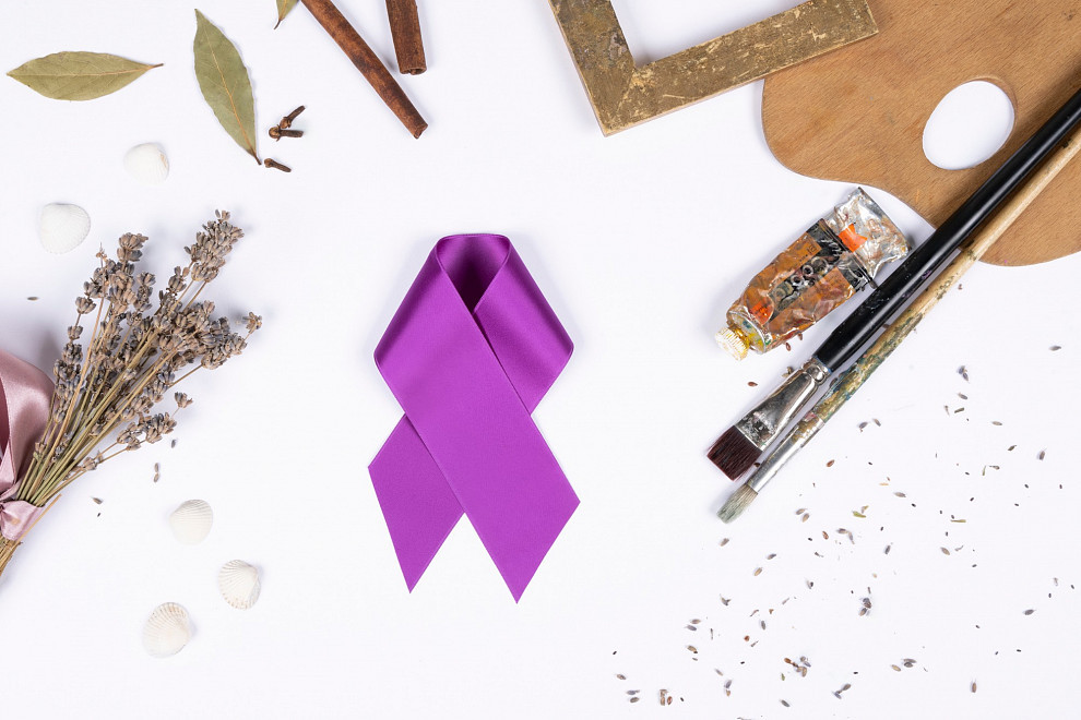 Eine violette Schleife auf weißem Hintergund, umgeben von dekorativen Accessoires wie Farben, Pinsel, einem Bilderrahmen und Lavendel | Foto © ALBERTINA Fotostudio