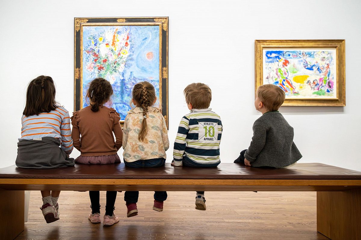 Fünf Kinder sitzen gereiht auf einer Bank in einer Ausstellung. Man sieht ihre Rücken und Hinterköpfe, vor Ihnen die Bilder Schläferin mit Blumen und der große Zirkus von Marc Chagall | Foto © ALBERTINA Fotostudio