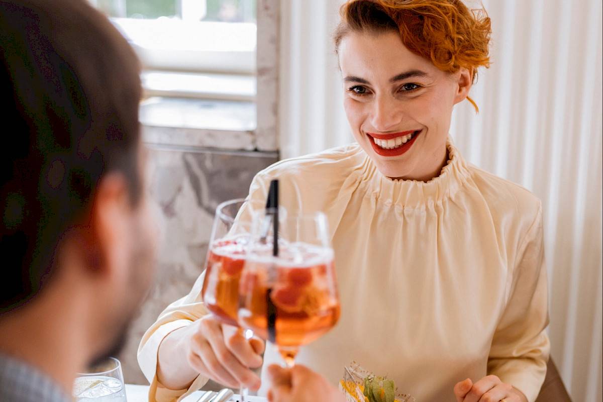Two people raise their glasses with Aperol. There is an orange cake in front of the smiling lady, seen from the front. | Photo © Marko Mestrovic