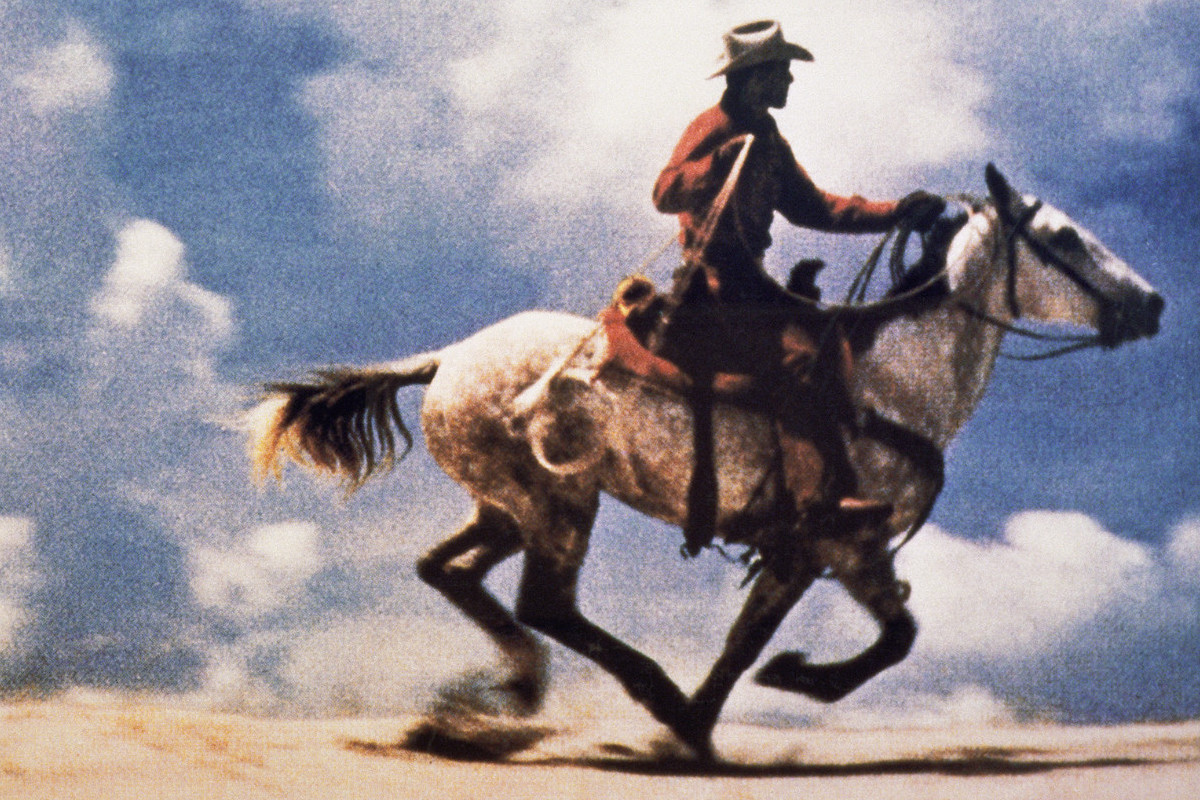 Color photograph of a cowboy riding a galloping horse in an open landscape; large-scale, horizontal composition.