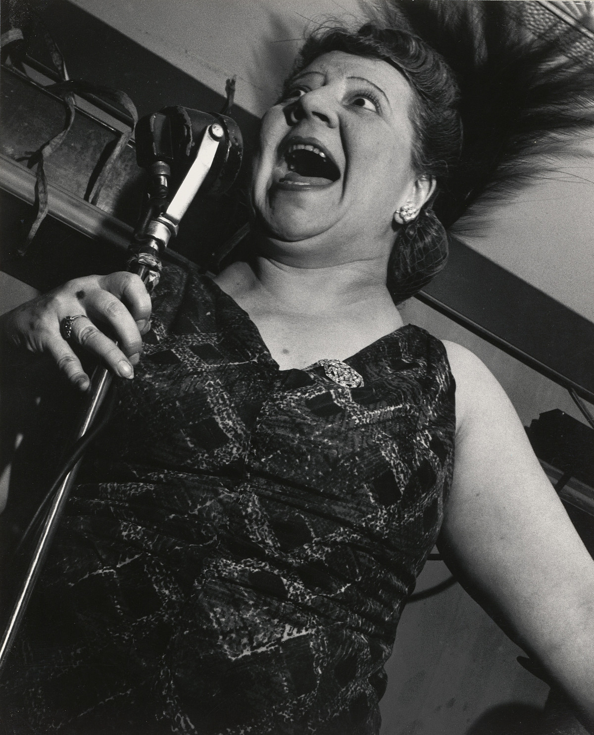 A woman in an evening dress holding a standing microphone up to her face. Her mouth is wide open.
