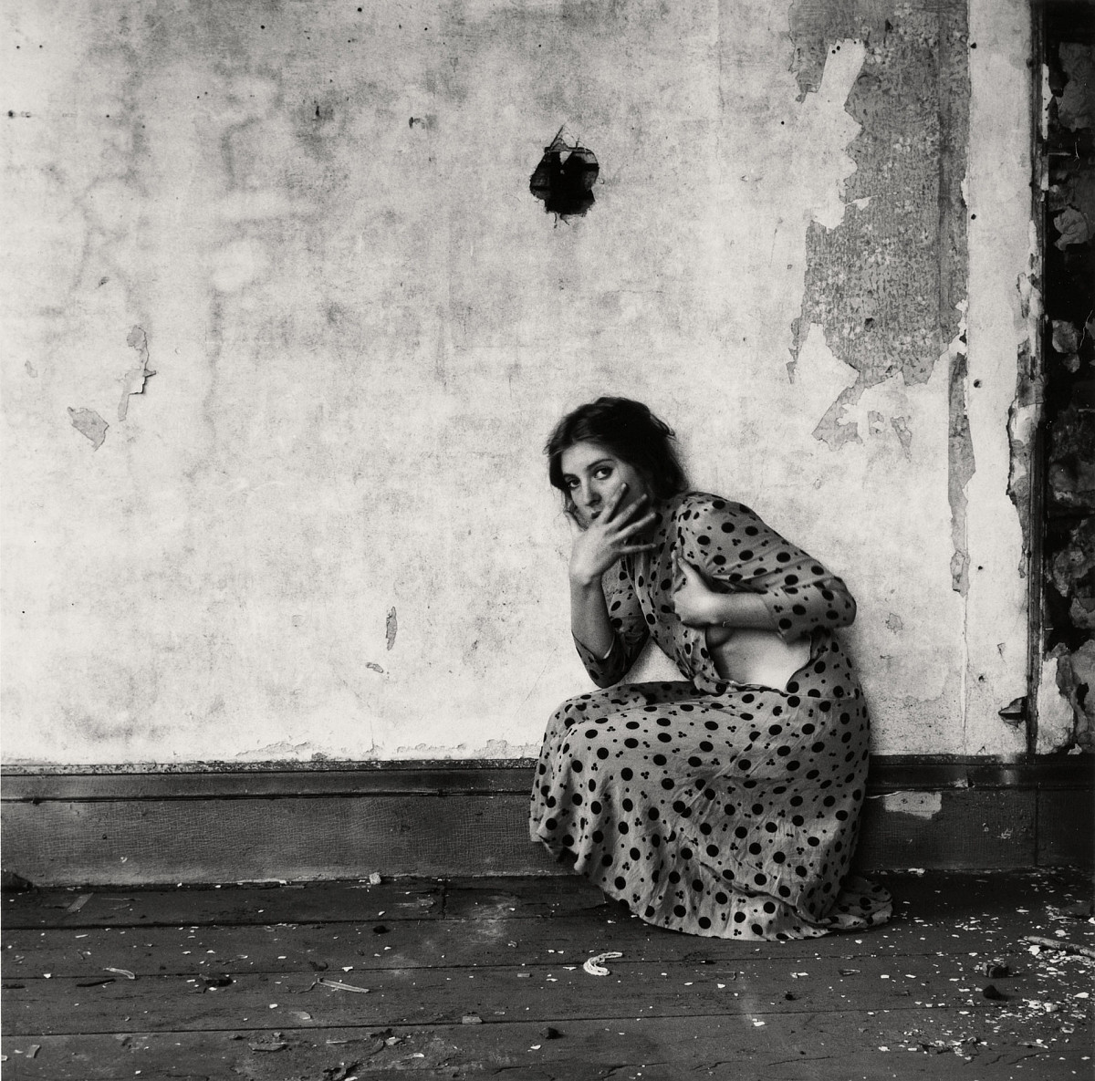 The artist kneels in front of a wall. She is wearing a long dress with a black polka dot pattern. She has turned to the side and is looking at the viewer. The zipper of the dress is attached to the side of the dress and is open. The wall and floor are very worn and cracked. There is even a round hole in the wall above the artist. The picture is a black and white photograph.