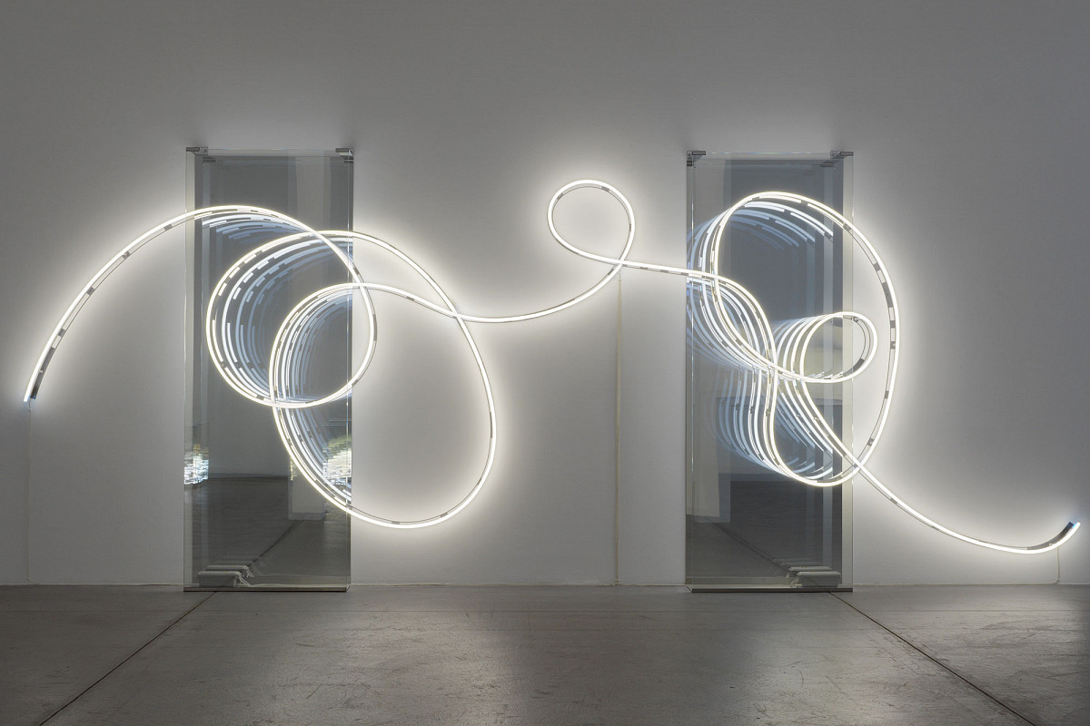 View into a dark exhibition room, illuminated only by a curved light installation in front of a mirror. | Brigitte Kowanz | Email 02.08.1984 03.08.1984, 2021 | ESTATE BRIGITTE KOWANZ © Estate Brigitte Kowanz / Bildrecht, Vienna 2025 | Photo: Peter Hoiss