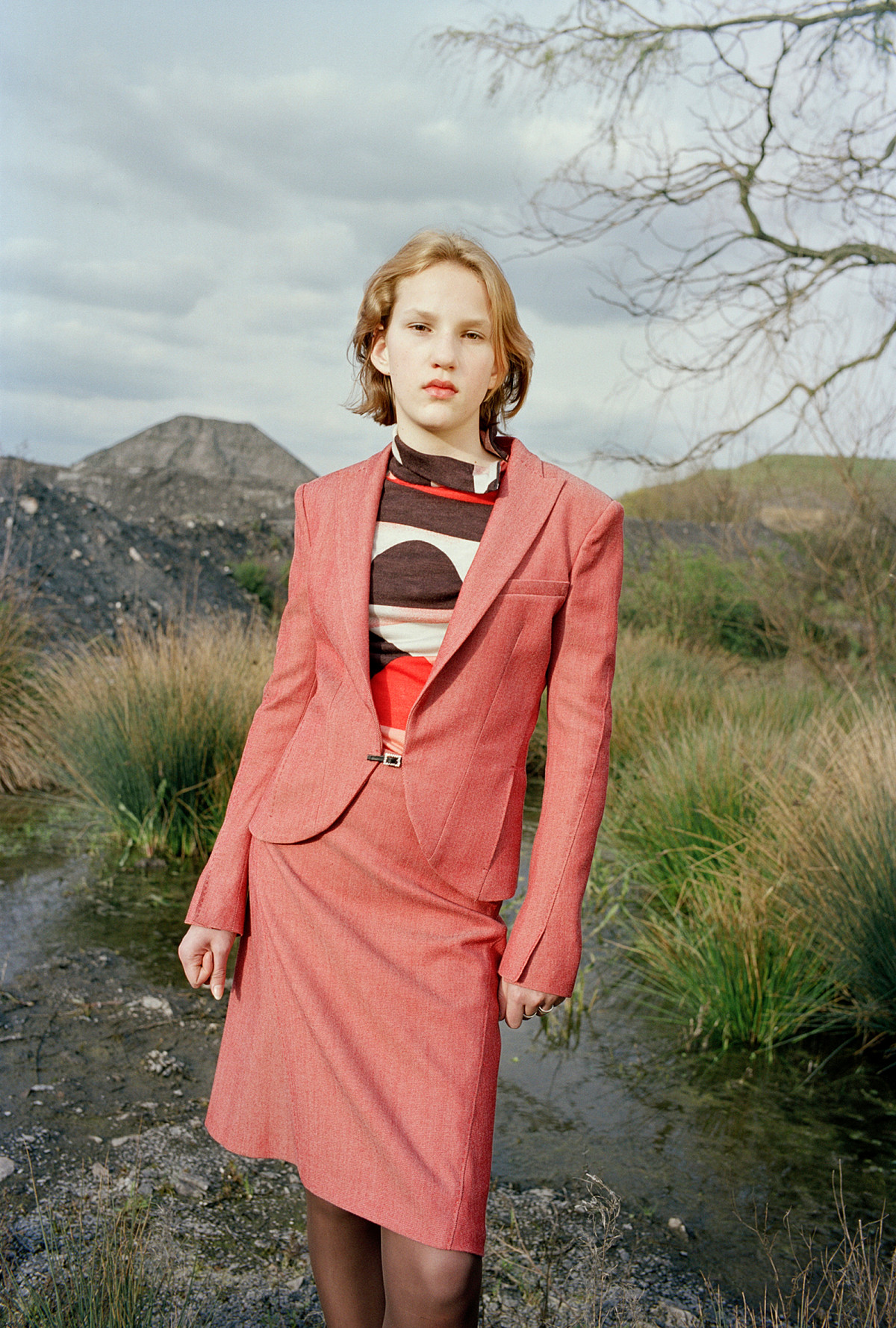 A woman stands in a barren landscape with a cloudy sky. She is wearing a pink skirt and blazer and has her hands clenched into fists.