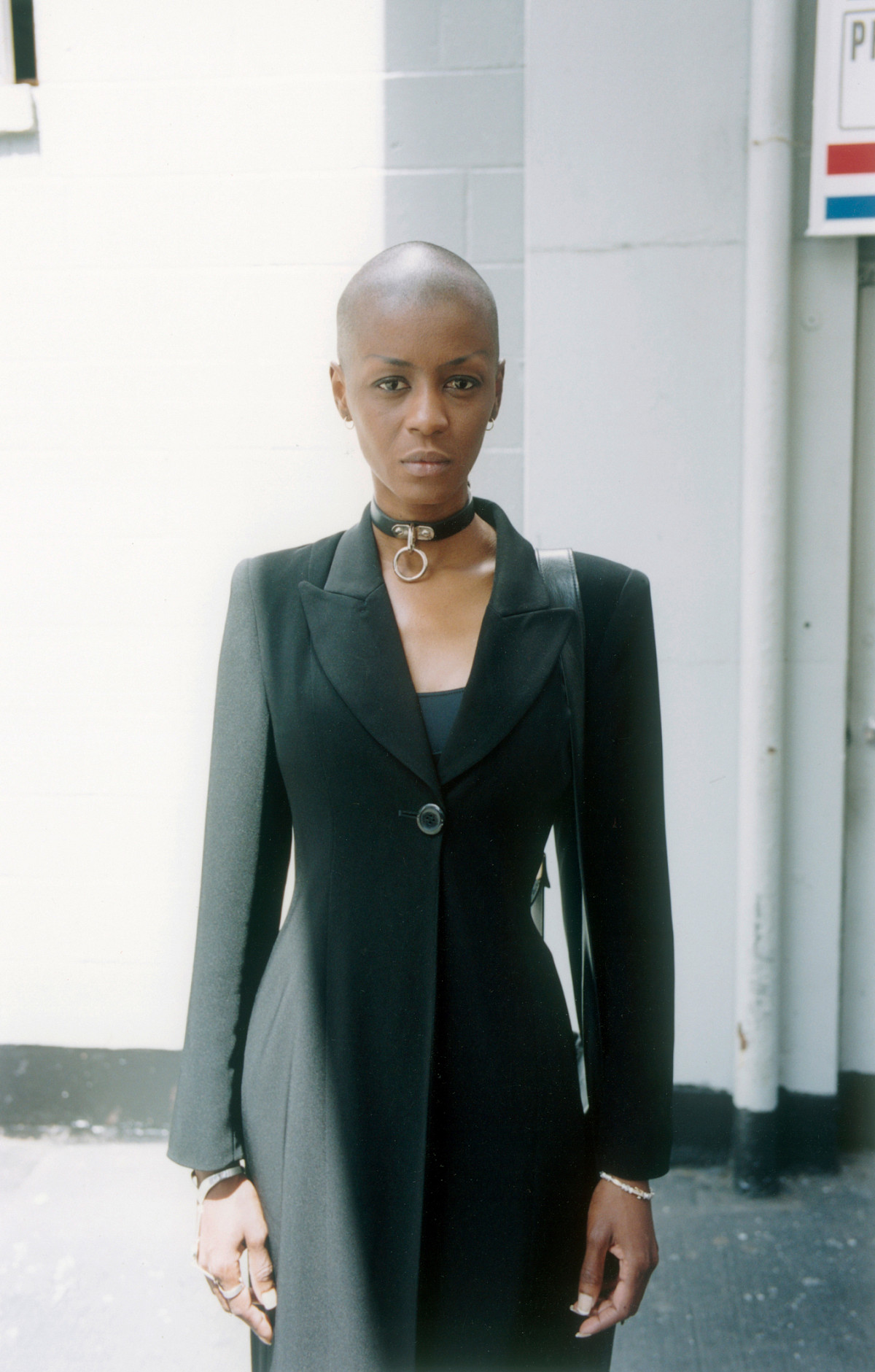 An African-American woman with a bald head, looking into the camera with a slight smile. She wears a black coat and a black leather band with a silver ring around her neck. A black bag hangs over her shoulder. A white building can be seen in the background.