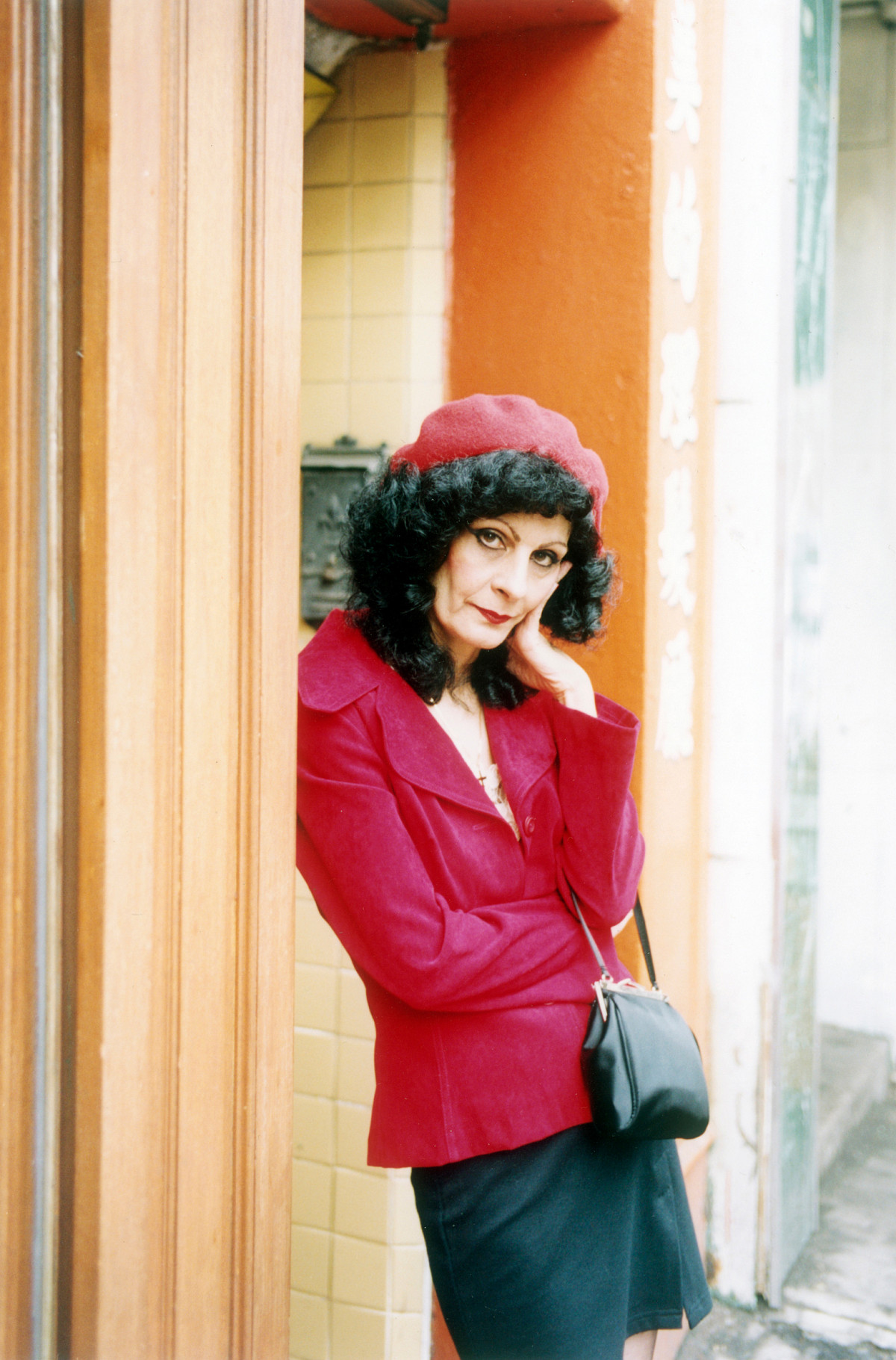 A woman with black curls and a red beret tilts her head and rests it on her hand. She looks into the camera and wears a red blazer jacket with a black miniskirt. A small black leather bag hangs in her hand. Her body is leaning against a door frame.