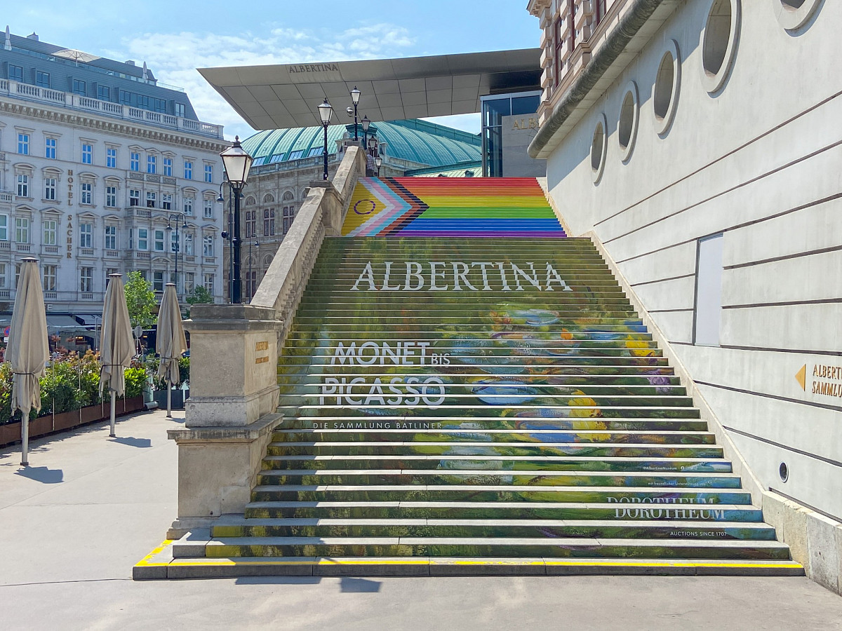 Die ALBERTINA-Treppe vor dem Gebäude mit der Beklebung. Motiv ist der Seerosenteich von Monet mit Aufschrift "Monet bis Picasso", Sponsorenlogos und dem Albertina-Logo. Darüber die Pride-Flagge (Intersex-Inclusive Pride Flag).