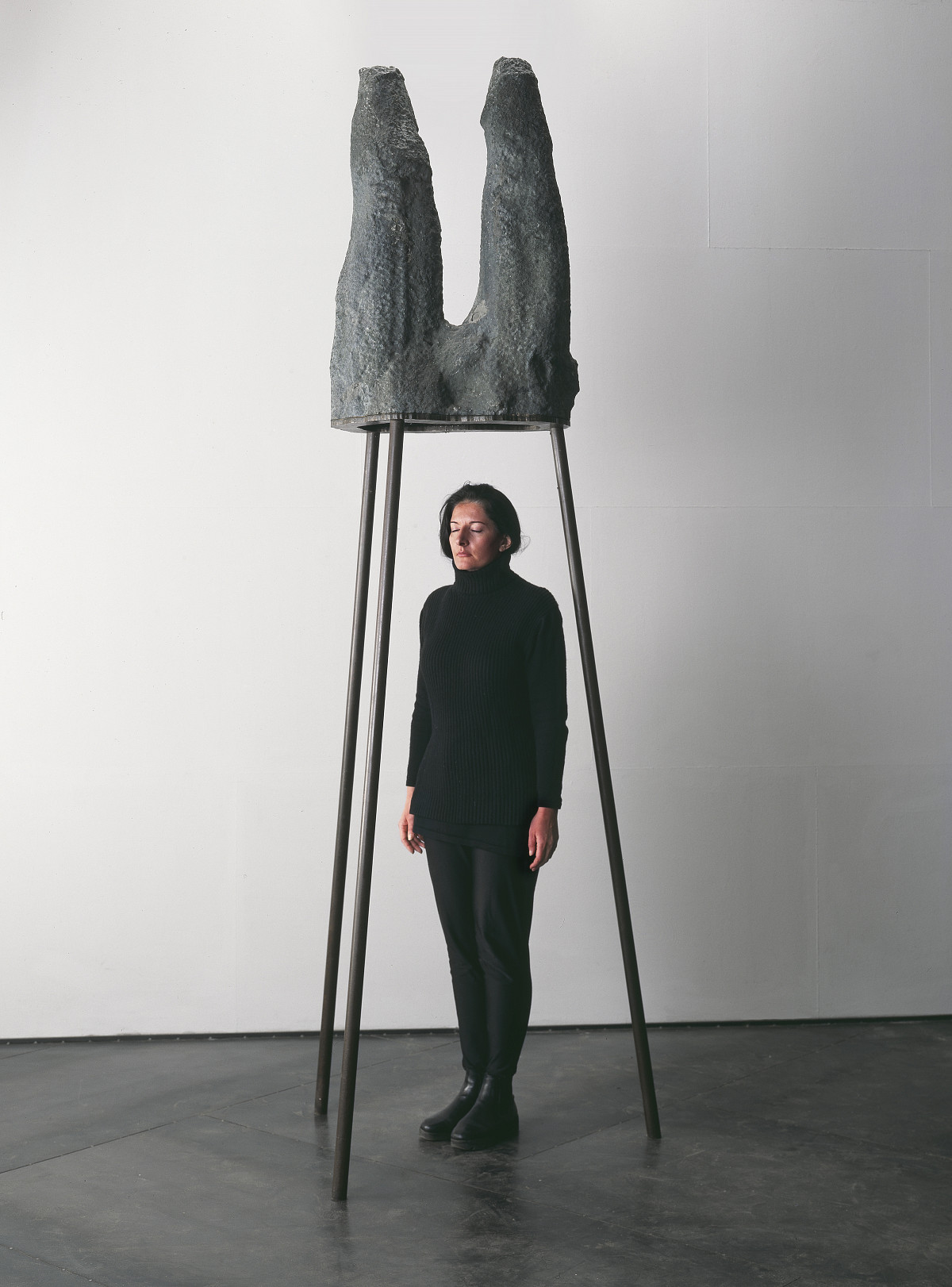 Marina Abramović stands upright under a large amethyst geode placed on three iron rods.