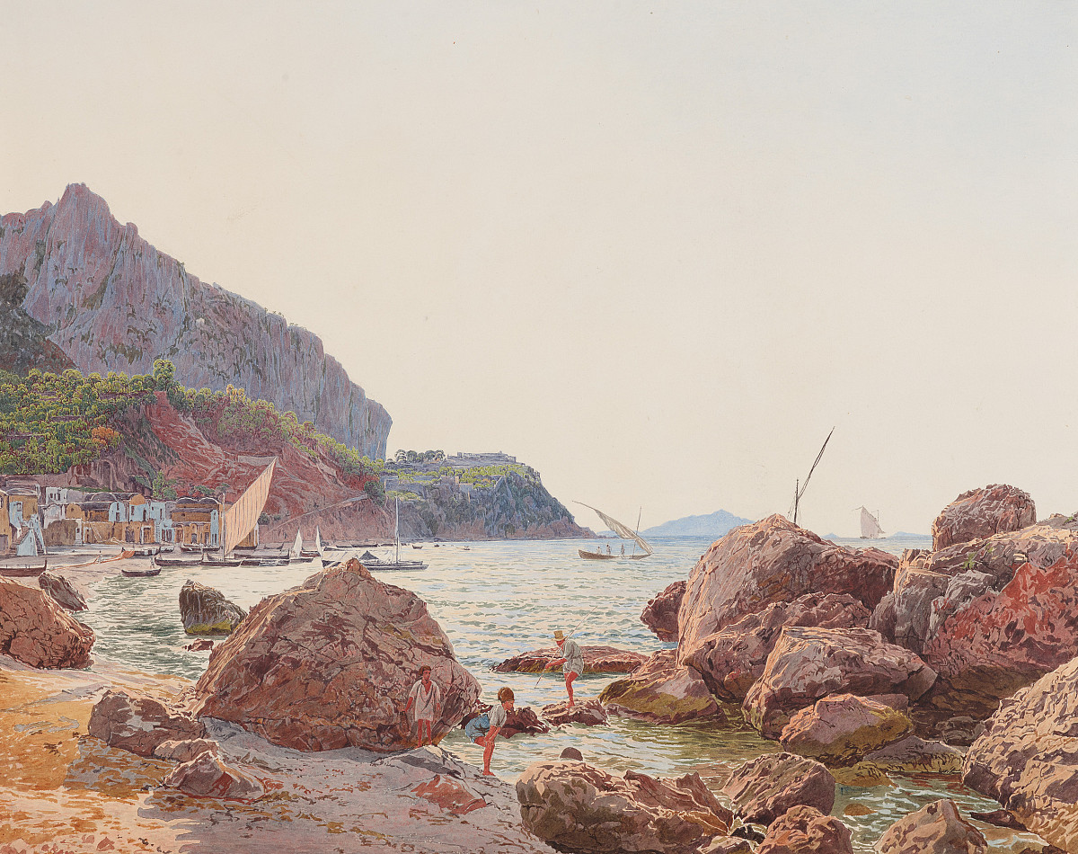 A beach in the foreground with the sea and large boulders on the beach and in the water. Two fishermen are standing by the rocks near the beach and a woman in a white blouse and blue trousers with a turban on her head is kneeling with her arms resting on her thighs looking at something on the beach. In the background is the harbor with small sailing ships and mountains.
