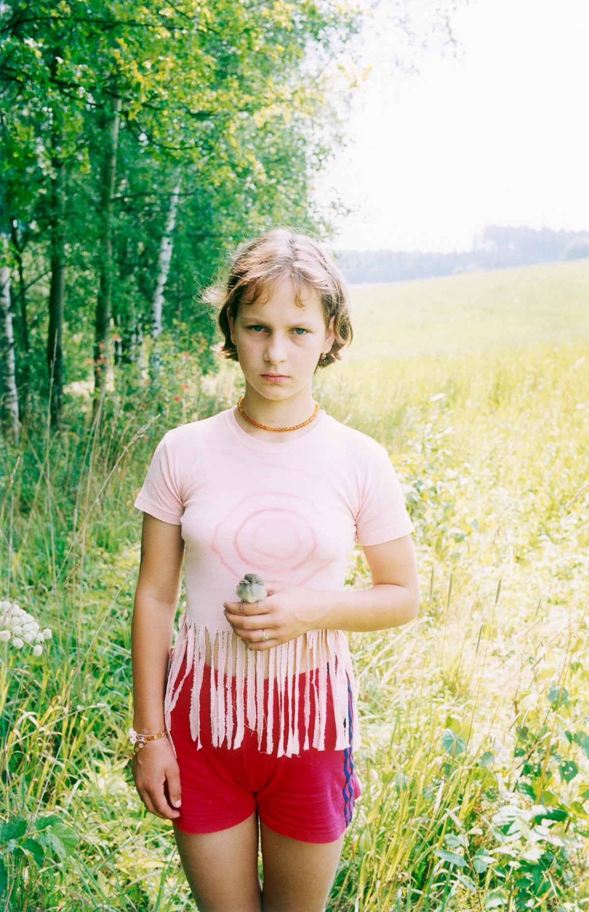 A young girl stands in a summer meadow in front of a forest and looks seriously into the camera. She is holding a flower in one hand.