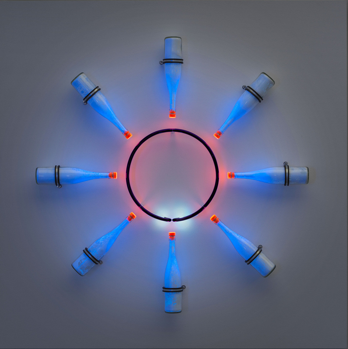 Installation of a black circle from which red light shines on eight glass bottles placed around it. These shine in a blue tone.