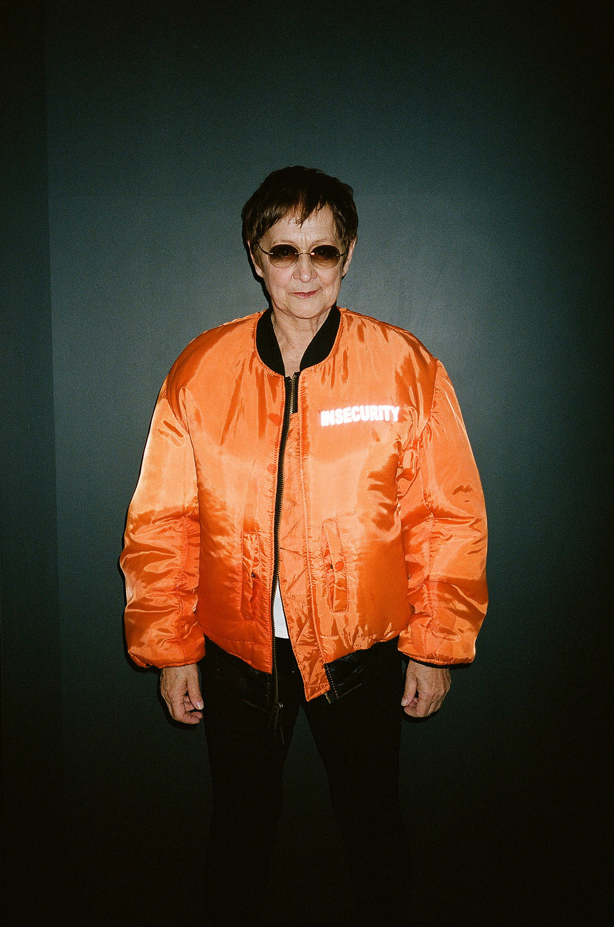 Portrait photo of the artist Brigitte Kowanz with orange bomber jacket and sunglasses.