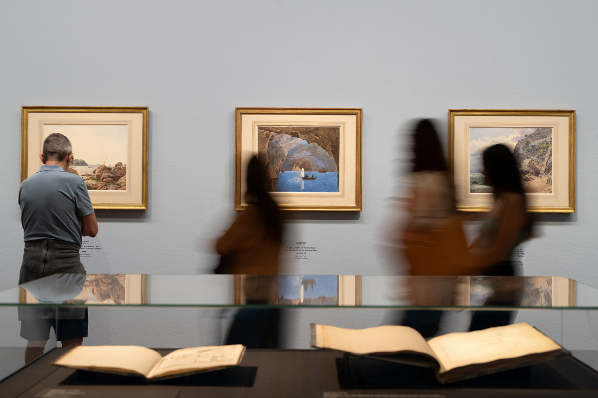 In der Ausstellungshalle: im Vordergrund ein Schaukasten mit aufgeschlagenen Büchern, im Hintergrund mehrere Gemälde an der Wand und Besucher:innen, die diese betrachten.