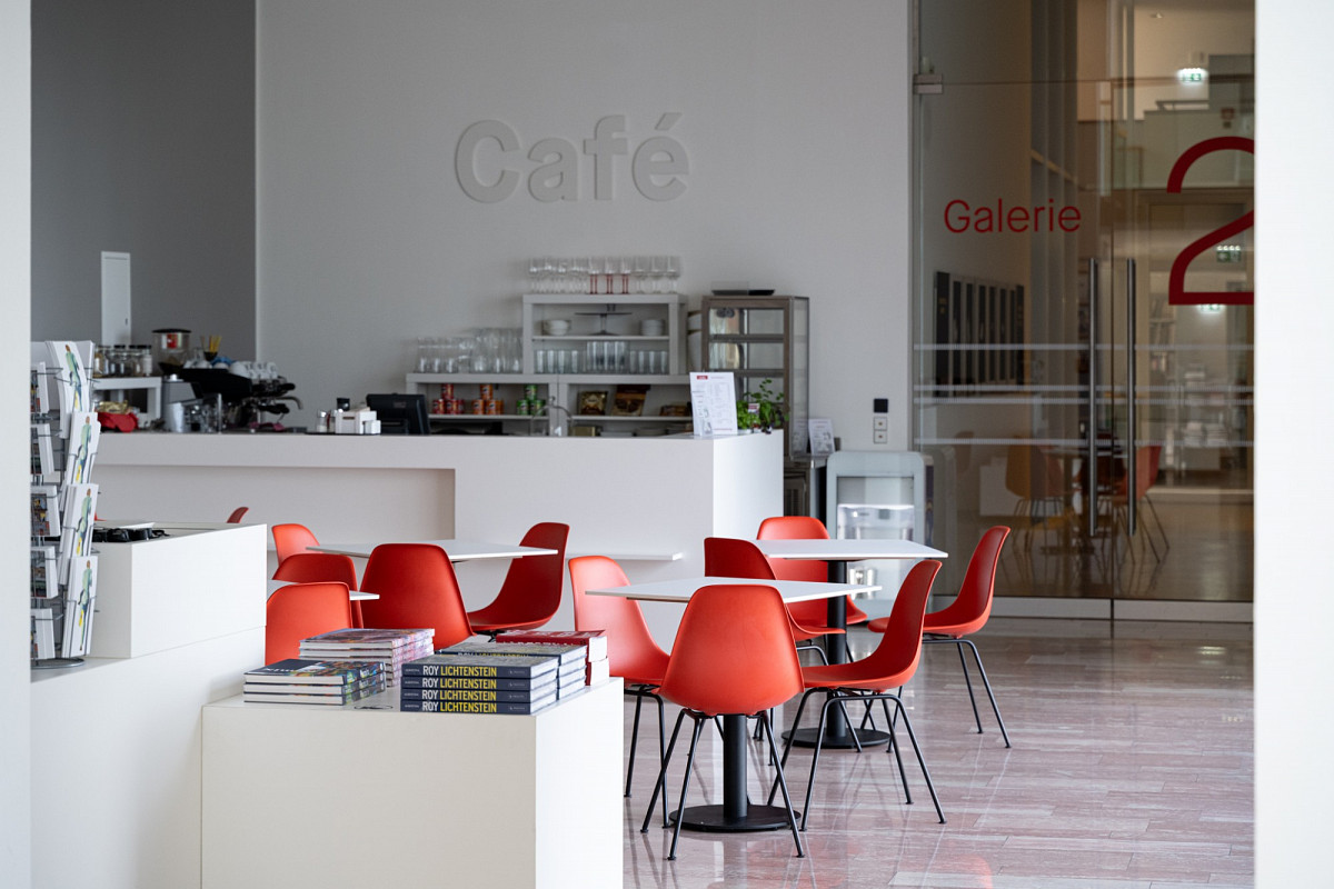 Café der Albertina Klosterneuburg: Weiße Tische mit orangenen Stühlen. Dahinter eine Theke und darüber die Aufschrift "Café" auf der Wand. Im Vordergrund ist ein Teil des Shops mit ausgestellten Katalogen zu sehen.