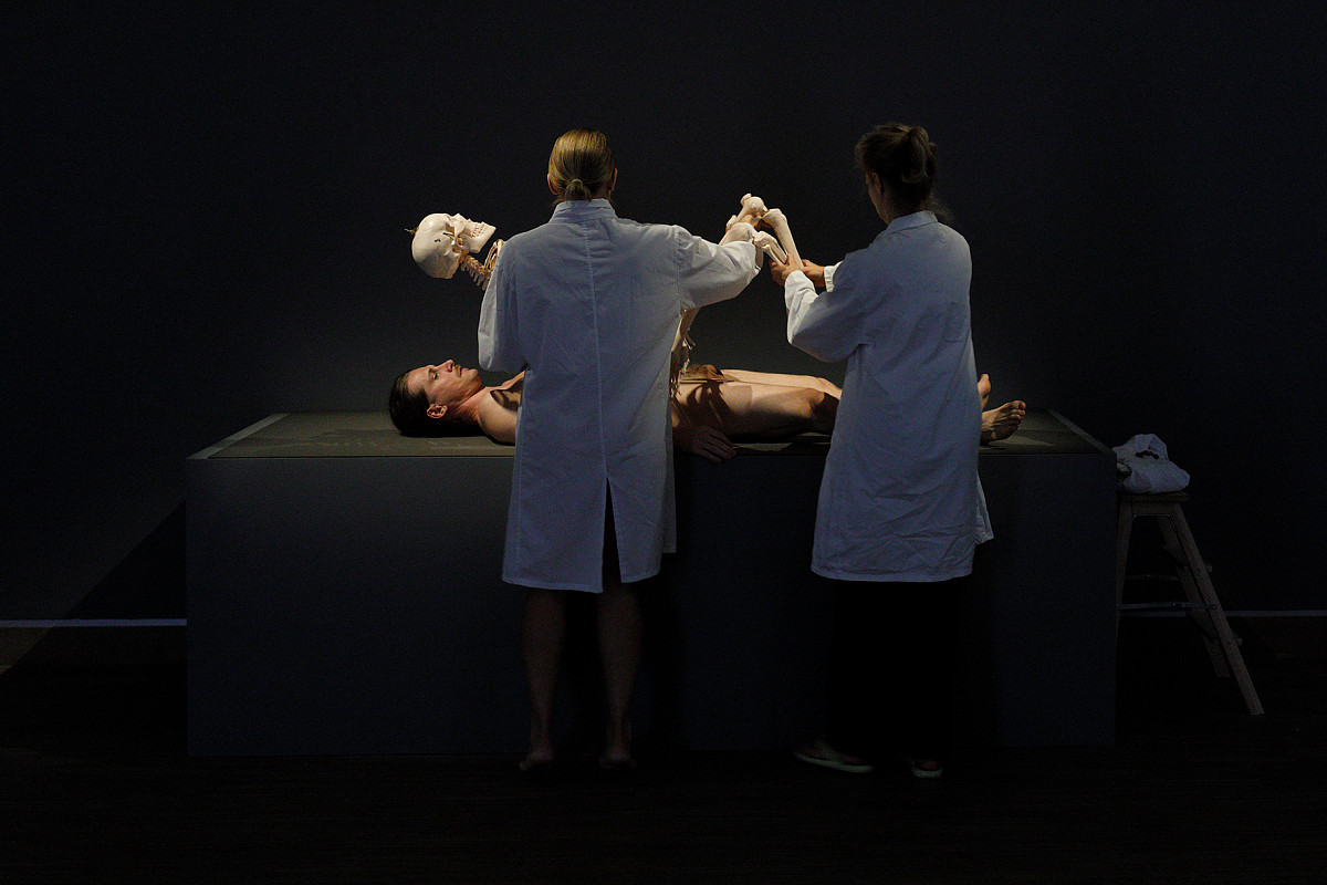 A naked man is lying on a table. In front of him are two people in white coats holding a human skeleton over his body.