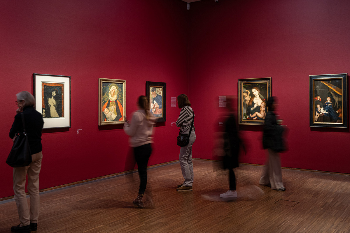 A red wall with several framed paintings next to each other, visitors standing in front of it.
