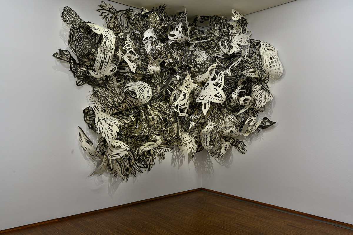 A corner of a room with a brown parquet floor and white wall. On the wall, in the corner, hangs a large installation of paper cut-outs in various patterns in black and white.