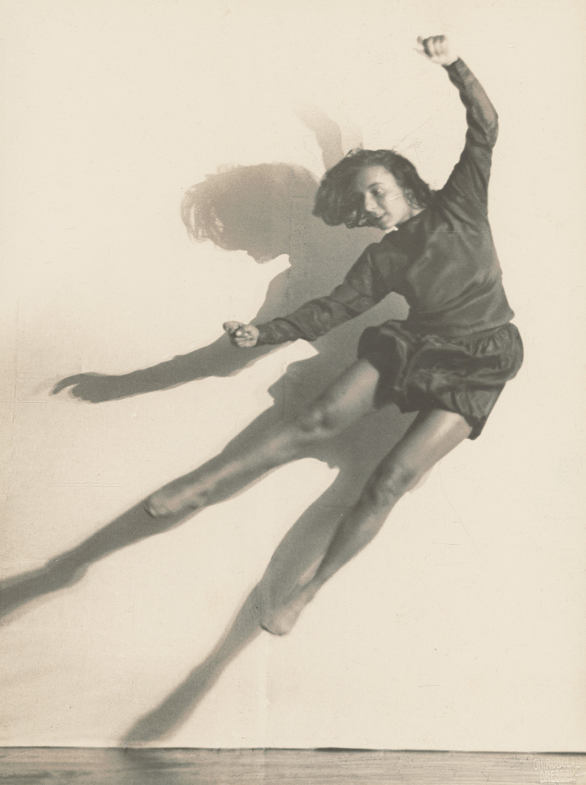 Photograph of dancer Gret Palucca jumping into the air, legs and arms stretched out wide. She is wearing a long-sleeved top and short, wavy pants.