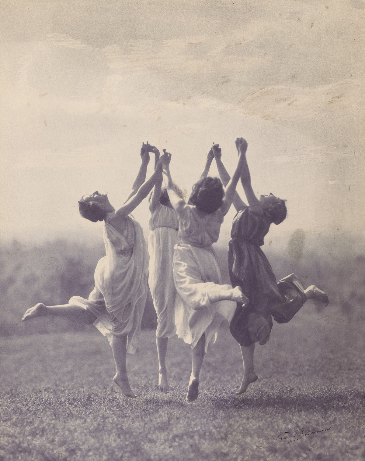 Photograph of four women in flowing dresses jumping on a meadow. They each stretch out one leg and hold hands. They stretch their hands toward the sky.