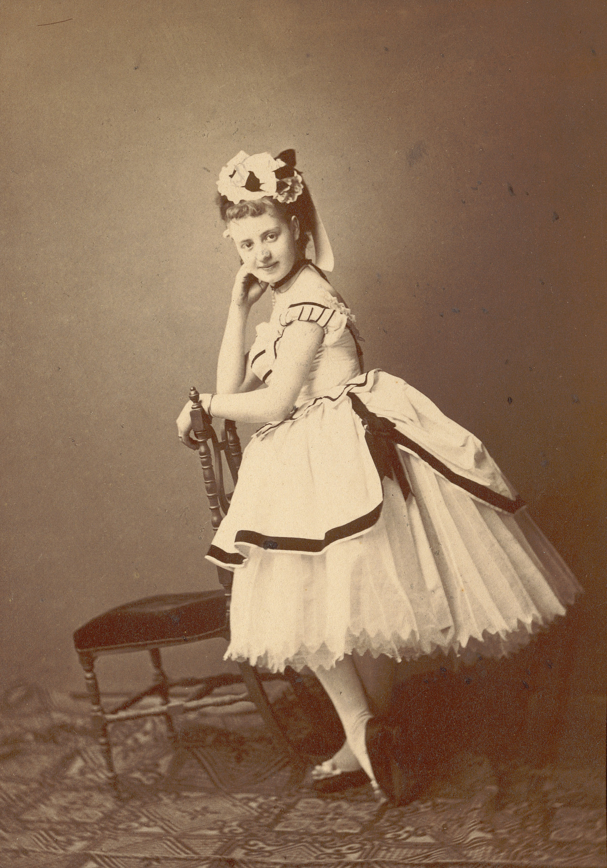 Photo of a dancer leaning against a chair, with a patterned carpet beneath her. She is wearing a floral headdress and a wide-skirted dance dress.