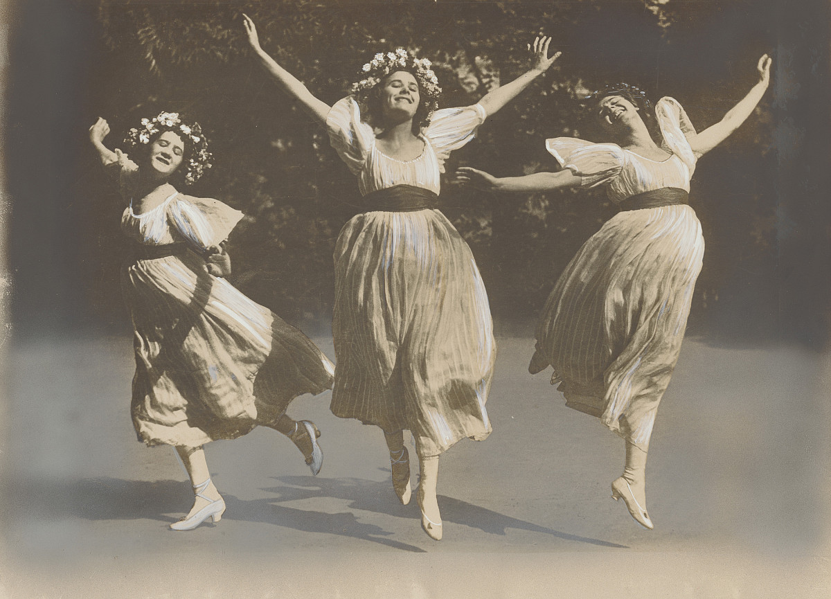 Photograph: Three women in flowing dresses with floral decorations and high heels jump and stretch their arms upward.