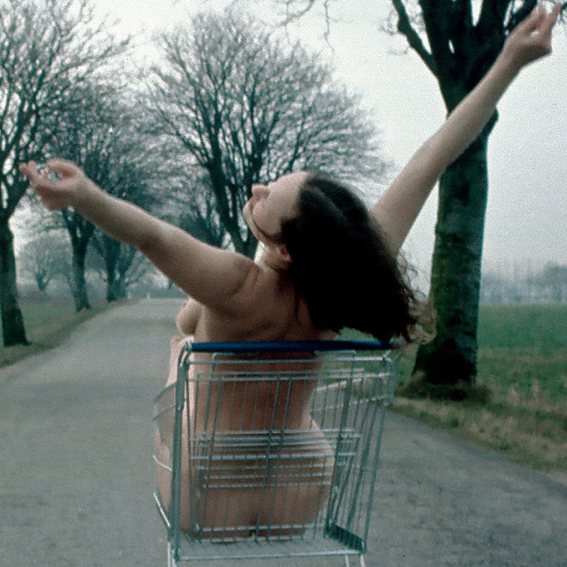Fotografie: Eine Frau sitzt nackt in einem Einkaufswagen und streckt die Arme in die Luft. Der Einkaufswagen steht auf einer leeren Landstraße. Links und rechts sind Felder und eine Allee mit kahlen Bäumen zu erkennen. Credit: Kirsten Justesen | Lunch, 1975/2018 | Courtesy SAMMLUNG VERBUND, Wien © Kirsten Justesen / Bildrecht, Wien 2026