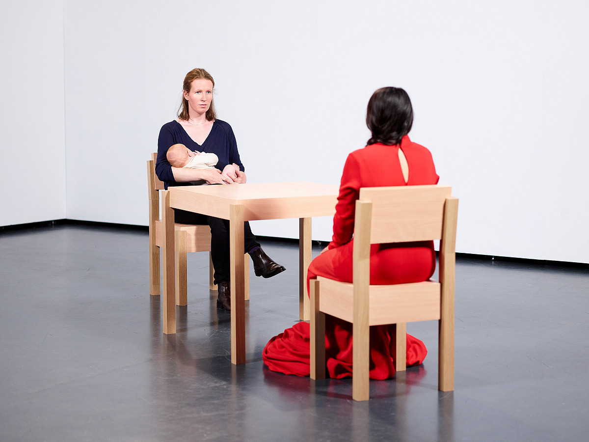 Two women sit opposite each other at a wooden table on wooden chairs. One woman holds a baby in her arms, while the other woman, with her back to the viewer, has black hair and wears a red dress.