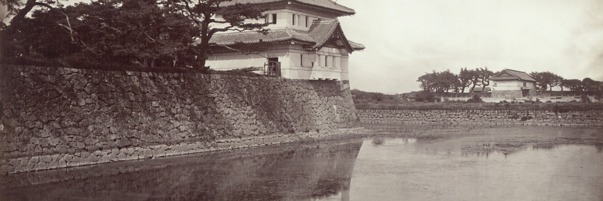 Schwarz-Weiß-Fotografie eines Wachgebäudes im japanischen Stil am Rande des Burggrabens und von Bäumen umgeben.