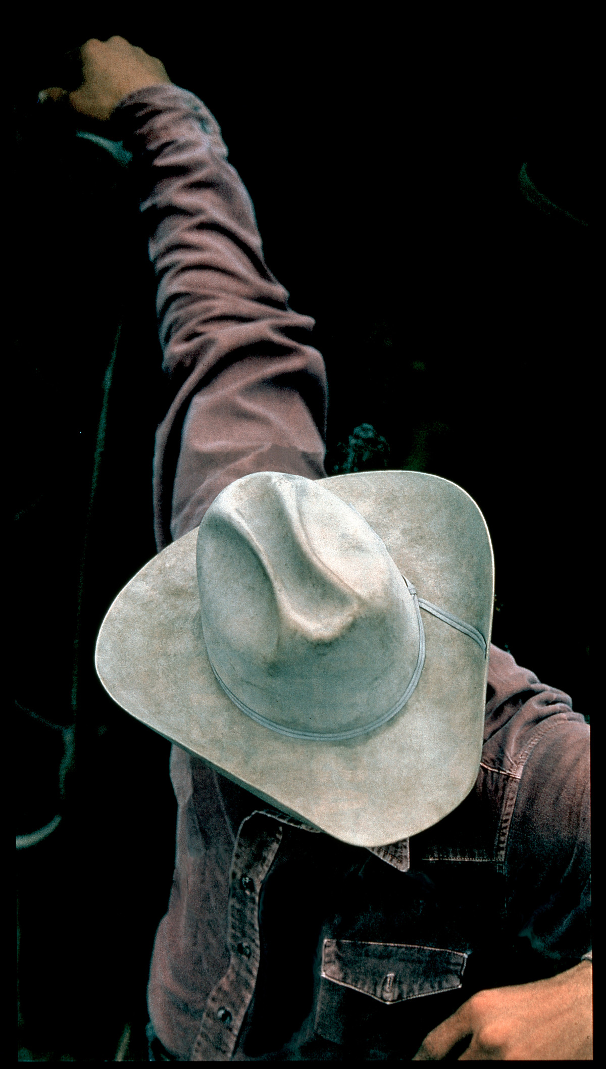 A photograph of a cowboy wearing a denim shirt and a hat, looking down with one arm raised.