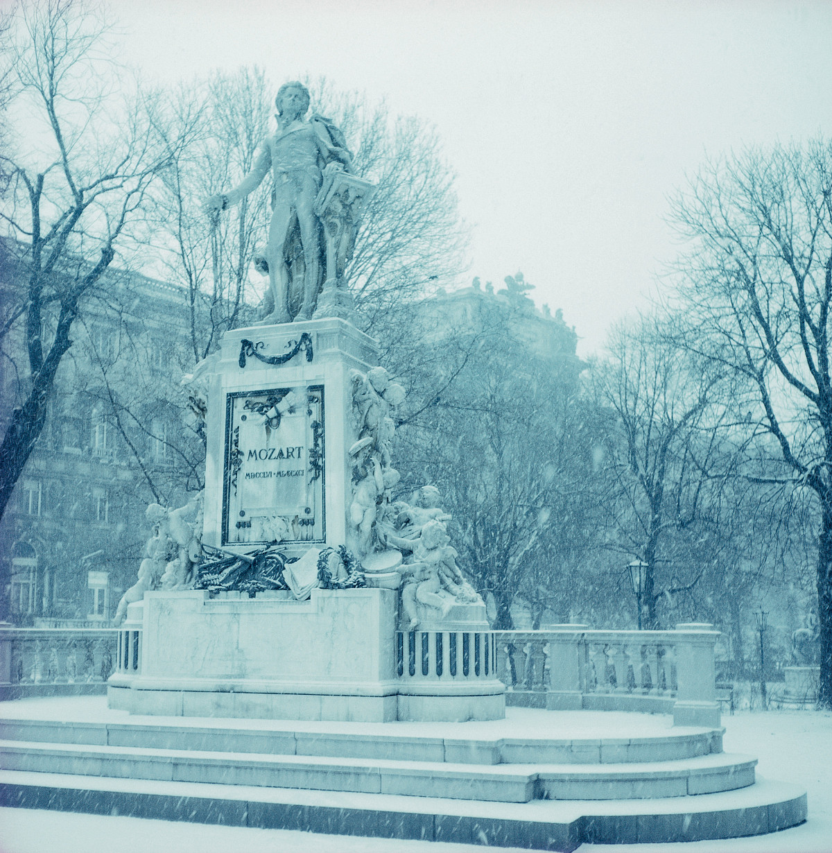 A photograph of a Mozart sculpture in a park. It is snowing, and a large building can be seen in the background.