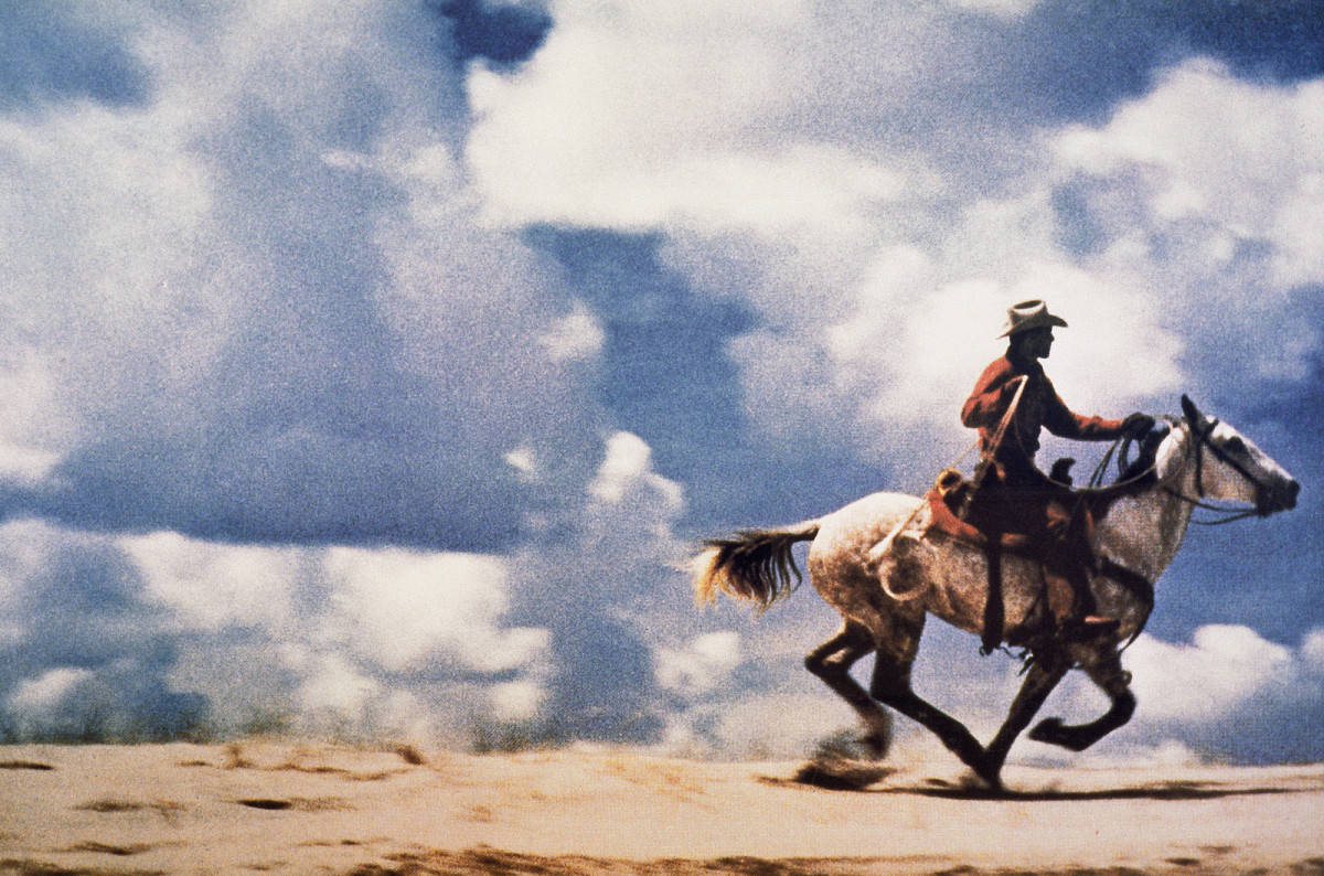 Color photograph of a cowboy riding a galloping horse in an open landscape; large-scale, horizontal composition.