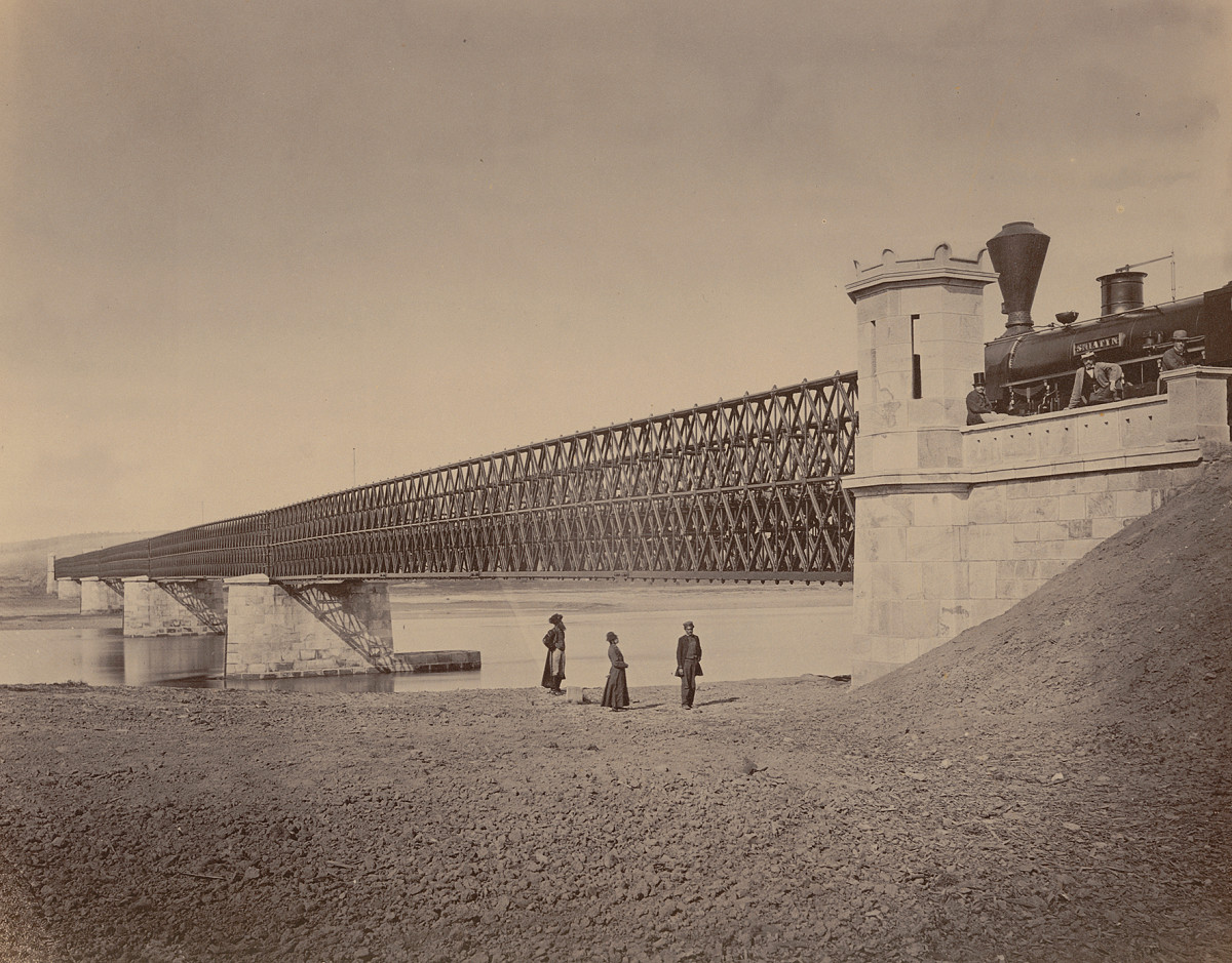 Photograph of a railway bridge spanning a river, with three people standing on the riverbank in the foreground.