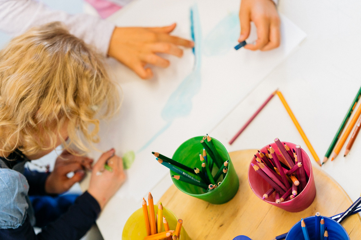 The focus is on several cups with different colored crayons. Next to it, a child paints with crayons, above you can see another child's hands. | Photo © Marko Mestrovic